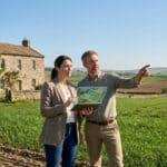 Professionals use a digital map on a tablet to discuss a large farm property with a stone house and green fields.