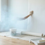 Gloved hand applies white fiberglass wallpaper to a wall, with paint tray and roller nearby. Ethereal mist suggests a healthy, breathable environment.
