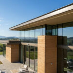 Modern house with extensive glass and warm rammed earth walls, set in a rolling green landscape under a bright blue sky with distant mountains.