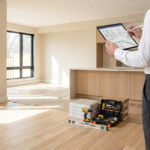 Personne en chemise blanche consulte des plans de rénovation sur tablette dans une pièce vide. Outils de chantier au sol.