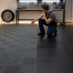 Un homme à genoux pointe du doigt deux types de dalles de sol de garage : une grise texturée (polypropylène) et une noire lisse (PVC).