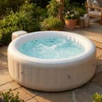 Spa gonflable blanc avec jets d'eau sur une terrasse en pierre, entouré de plantes et de fleurs. Lumière dorée, mobilier de jardin en fond.