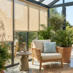 Véranda lumineuse avec fauteuil, plantes et stores beiges filtrant le soleil estival.