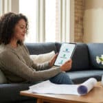 Une femme souriante consulte une application de rénovation sur tablette assise dans un salon lumineux.